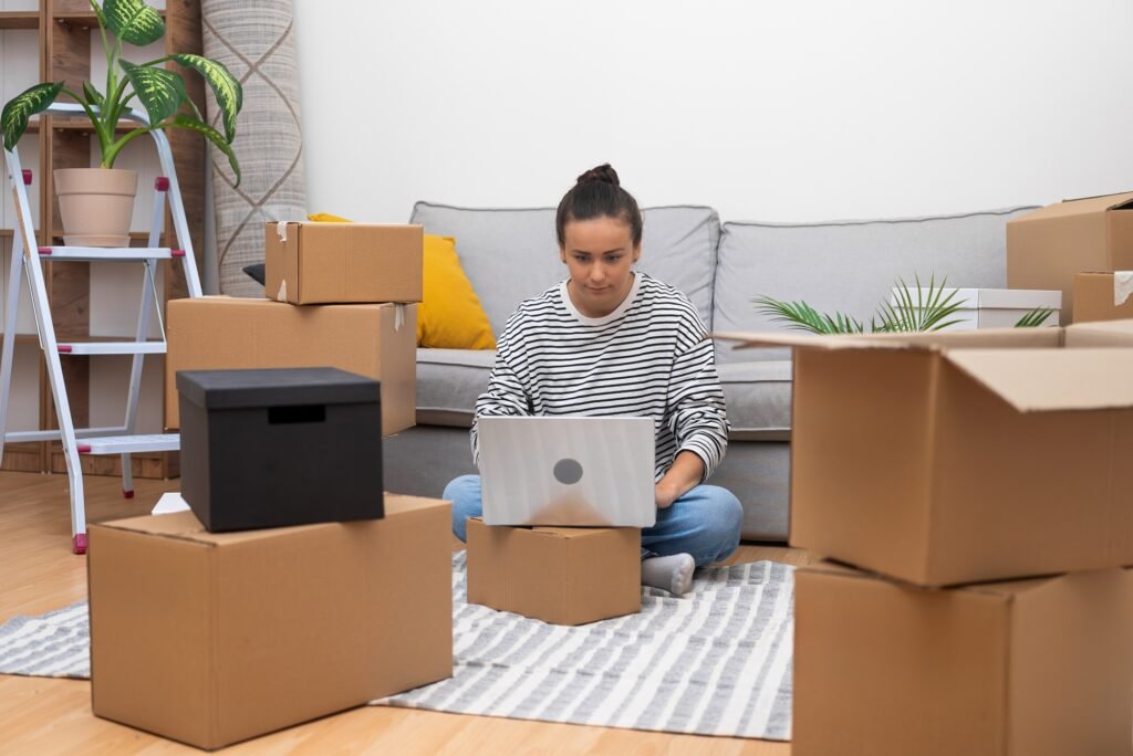 woman surrounded by boxes, uses a laptop to plan home decor, shop online, or book removal services