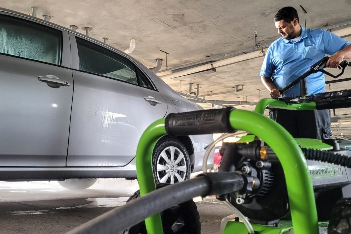 Millennial maintenance man at condo uses digital power washer to clean a Toyota corolla in garage.